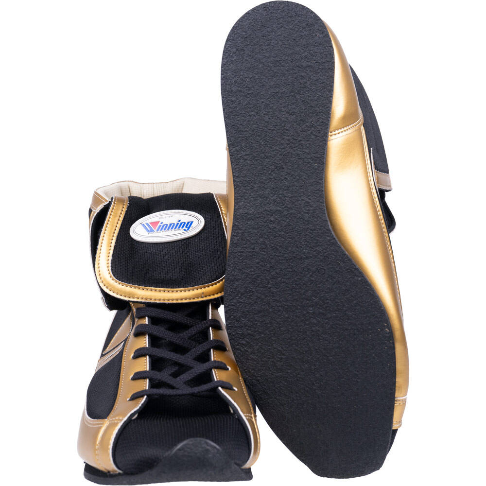A pair of boxing shoes in black and metallic gold, with one shoe standing upright to show the side profile and logo, and the other lying on its side to display the textured black outsole.